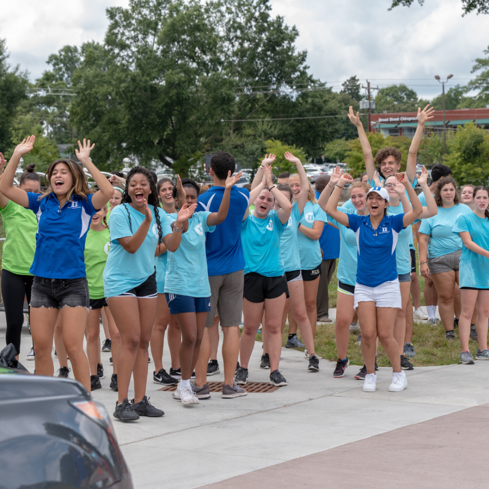 students in multi-colored tshirts cheer on new students moving in