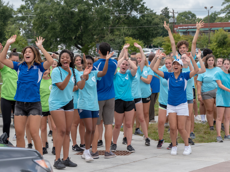 students in multi-colored tshirts cheer on new students moving in