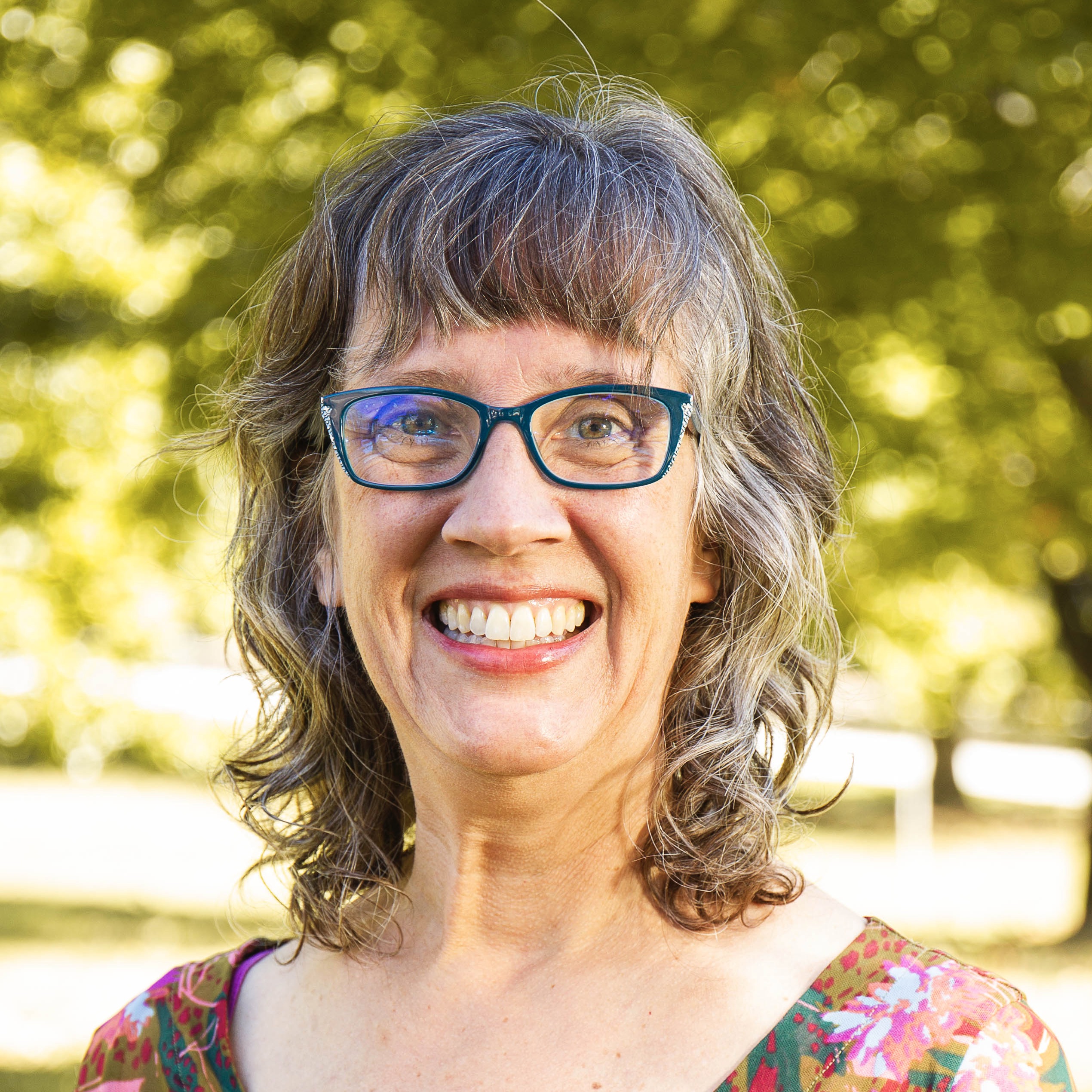 A white woman with light brown hair and blue glasses wearing a purple, patterned top