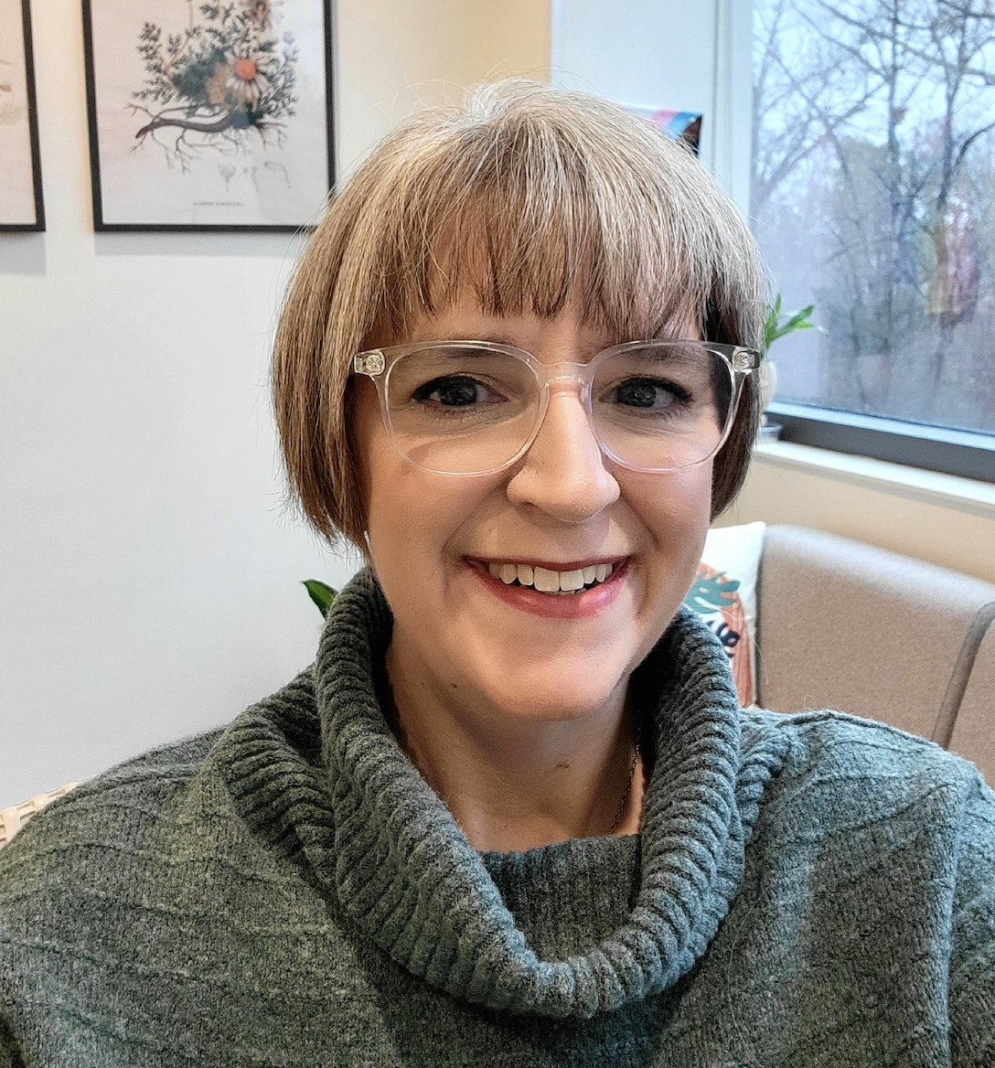 Photo of Amy Johndro–smiling white woman with short, light-colored hair wearing clear glasses and a grey-green cowl sweater