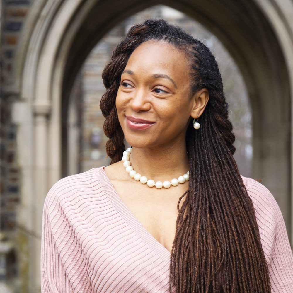 photo of an African American women (Women's Center director Krystal George) with a pink shirt and pearls standing in front of the archways of the Duke chapel