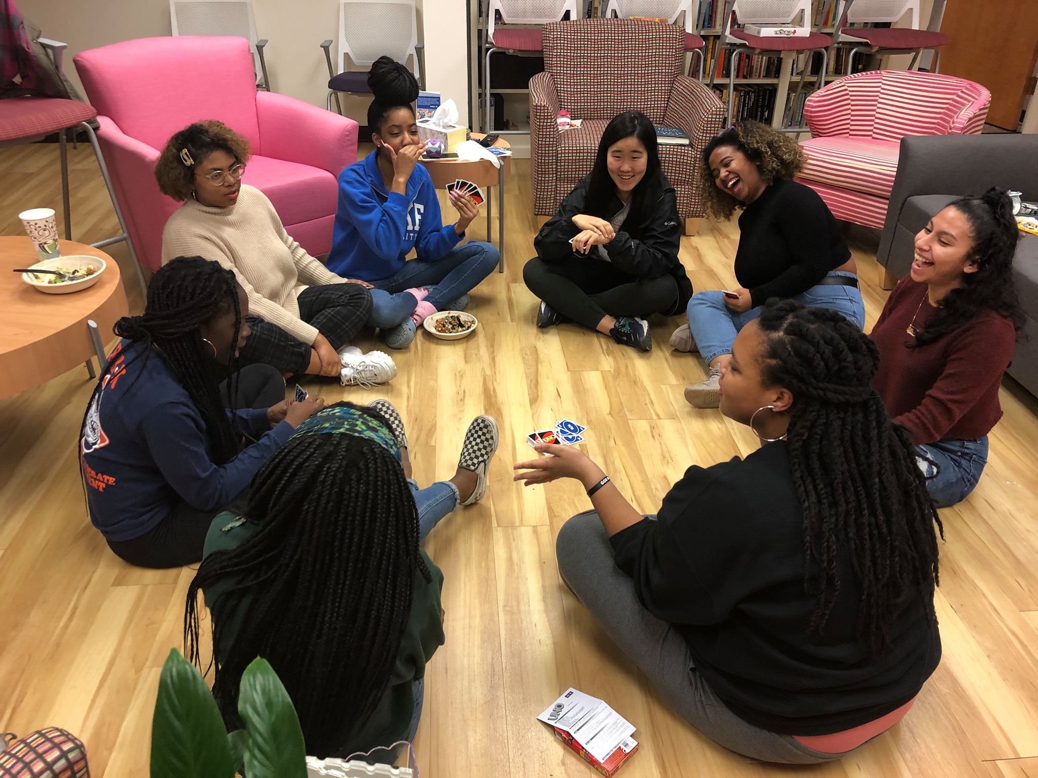 a group of women/femme-identified folks of medium to dark skin tones sit in a circle on a light wooden floor playing a card game while laughing and smiling jovially