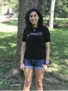 a woman with an olive-skin tone stands outdoors with her back to a tree in the middle of a sun-dappled grassy field; she is wearing denim shorts and a black t-shirt with the words "still feminist" printed on it in white text; she stands in the center of the photo, depicted from her knees up with her arms by her side smiling at the camera