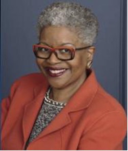 a dark-skinned woman with greying hair, red glasses, and a red suit jacket smiles at the camera with burgundy lipstick