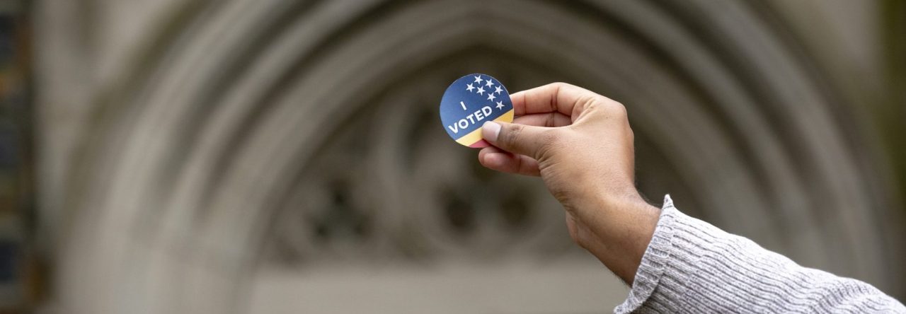 Photo of a student's hand holding a blue "I Voted" sticker in front of a Duke arch